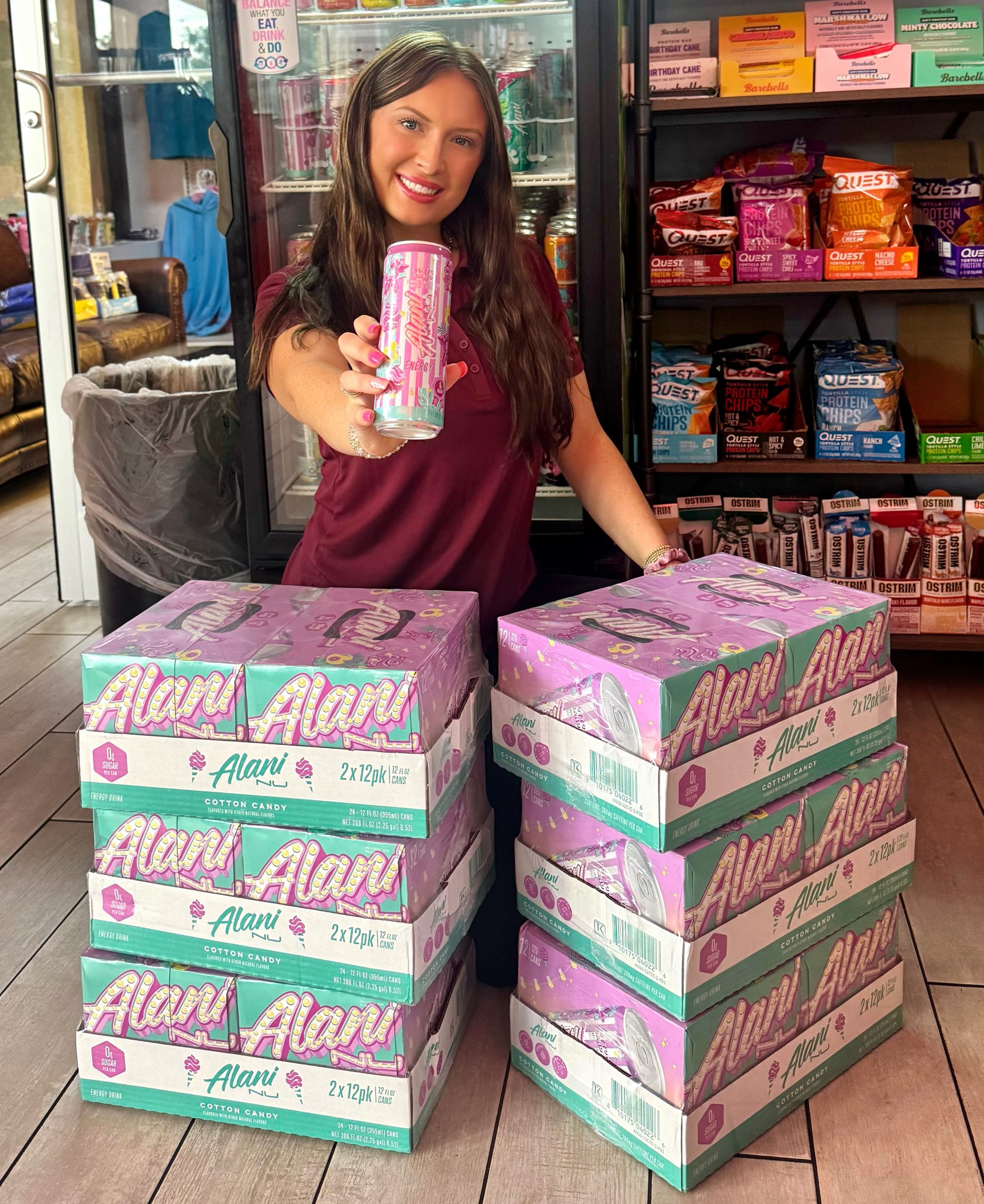 Girl posing in front of a stack of "Alani Nu" Energy Drinks. She is holding one pink can towards the camera, smiling.